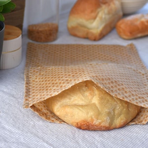 May include: A loaf of fresh, golden-brown bread partially wrapped in a tan, patterned cloth. Other loaves of bread are visible in the background, along with a jar of seeds and a small container with a wooden lid. The scene suggests a bakery or kitchen setting.