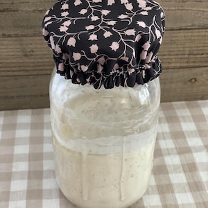 May include: A glass jar with a white sourdough starter inside, covered with a black and pink floral fabric lid.