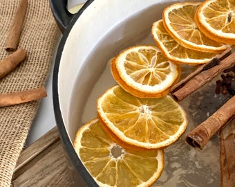May include: A black and white enamel pot filled with water, orange slices, cinnamon sticks, cloves, and apple slices. The pot sits on a wooden cutting board next to a burlap sack. The ingredients suggest a warm, spiced beverage or potpourri.