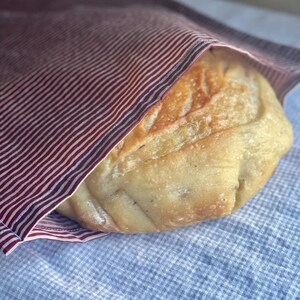May include: A loaf of freshly baked bread wrapped in a burgundy and white striped cloth. The bread has a golden-brown crust and is resting on a blue and white checkered cloth.