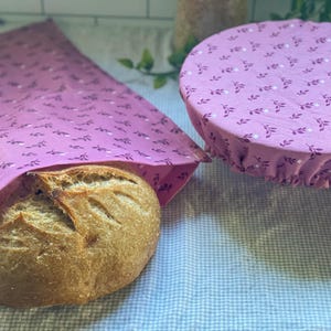 May include: A pink fabric bread bag with a small dark purple leaf and white dot pattern containing a round loaf of crusty bread, alongside a matching round elasticized fabric bowl cover with the same floral print pattern.
