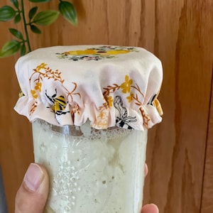 May include: A clear glass jar filled with a thick, off-white sourdough starter, topped with a fabric cover. The fabric is light pink and features a repeating pattern of yellow and black bees, small yellow flowers, and green leafy vines.