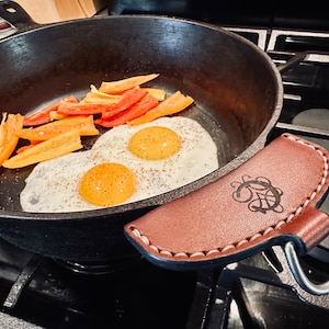 May include: Two fried eggs with pepper seasoning in a black cast iron skillet with orange and yellow bell pepper slices. A brown leather handle cover with a decorative design is on the handle of the skillet.