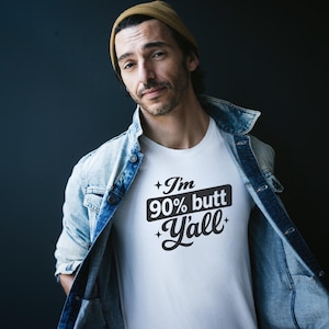 Könnte beinhalten: Weißes T-Shirt mit dem Text "I'm 90% butt Y'all" in einem schwarzen Rechteck. Die Person auf dem Bild trägt eine hellblaue Jeansjacke und eine beige Mütze. Der Hintergrund ist dunkelblau.