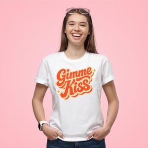 May include: White t-shirt with the text "Gimme Kiss" in orange script with yellow star accents. The shirt is worn by a person with sunglasses on their head and a watch on their wrist. The background is a solid pink color.