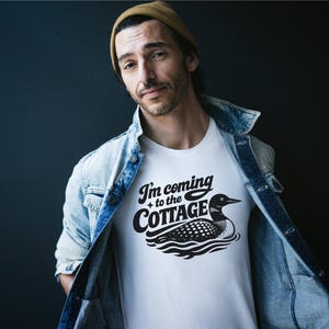 May include: White t-shirt with the text "I'm coming to the Cottage" and a black and white loon graphic. The person is wearing a light blue denim jacket and a tan beanie.