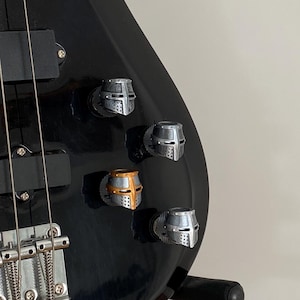 Peut inclure: Une guitare basse électrique noire avec une touche en bois et des mécaniques argentées. La guitare a un chevalet argenté et un ensemble de boutons en forme de casque de chevalier argentés.