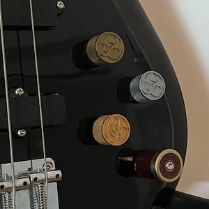 May include: Close-up of a black bass guitar with gold, silver, and bronze knobs. The bass guitar is on a black stand, and the body is angled towards the viewer. The knobs have a unique design.