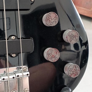 Könnte beinhalten: Nahaufnahme einer schwarzen E-Bassgitarre. Die Gitarre hat silberne Stimmwirbel und Saiten, mit vier Bedienknöpfen, die komplizierte Designs zeigen. Der Korpus ist glänzend schwarz.