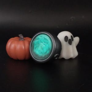 Könnte beinhalten: Eine Halloween-Arrangement mit einem kleinen orangefarbenen Kürbis, einem silbernen Ring mit einem leuchtenden türkisfarbenen Stein und einer weißen Geisterfigur. Der Ring hat ein dunkles Metallband. Die Objekte sind vor einem dunklen Hintergrund platziert.