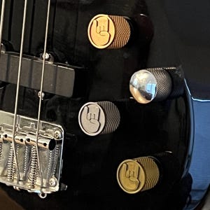 以下が含まれることがあります： 銀色の弦とハードウェアを備えた黒いエレクトリックベースギターのクローズアップ。ギターには、金と銀の手のジェスチャーデザインの3つのコントロールノブがあります。楽器は黒いスタンドに置かれています。