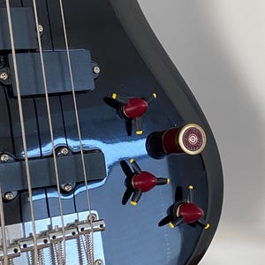 Puede incluir: Un bajo eléctrico negro con un acabado brillante. La guitarra tiene cuatro cuerdas, pastillas negras y perillas de control rojas y amarillas. El puente y las clavijas de afinación son plateados. El instrumento está sobre un soporte negro.