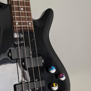 Puede incluir: Un bajo eléctrico negro con un diapasón de madera y herrajes cromados. El bajo tiene cuatro cuerdas y está decorado con cinco pequeñas pegatinas redondas de colores.