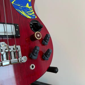 Könnte beinhalten: Eine rote elektrische Bassgitarre mit einer gelb-blauen Grafik auf dem Korpus. Die Gitarre hat schwarze Knöpfe, einen Chromsteg und schwarze Stimmwirbel. Die Knöpfe sind wie Moai-Statuen geformt.