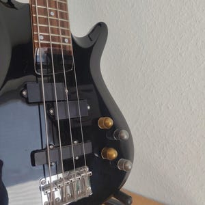 Pode incluir: Um baixo elétrico preto com acabamento brilhante. O instrumento apresenta uma escala de madeira, ferragens cromadas e botões de controle dourados e prateados. O baixo está apoiado em um suporte preto contra uma parede branca.