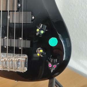 May include: A glossy black electric bass guitar body featuring three custom control knobs shaped like plague doctor masks wearing top hats, with neon yellow, pink, and green accents, alongside a circular teal knob with a graphic design.