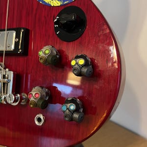 Könnte beinhalten: Eine rote E-Gitarre mit vier individuellen Knöpfen in Form von Gasmasken. Die Gasmaskenknöpfe haben verschiedenfarbige Gläser: grün, gelb, rot und blau. Die Gitarre hat einen schwarzen Lautstärkeregler und einen Aufkleber oben.