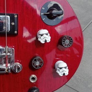 Könnte beinhalten: Ein roter E-Bass mit schwarzen Knöpfen und einem schwarzen Schlagbrett. Die Knöpfe haben Köpfe von Star Wars Stormtrooper-Charakteren.