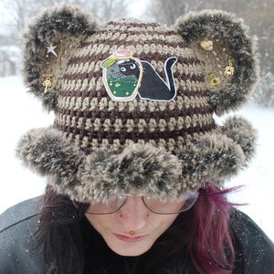 May include: A brown and tan crocheted hat with faux fur trim and ear-like accents. The hat features a patch of a black cat in a jar and is adorned with gold-colored charms. The hat is worn in a snowy outdoor setting.