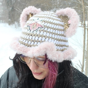 May include: A handmade crochet hat with a white and tan striped pattern, adorned with pink faux fur trim and ear-like accents. A moth applique is centered on the hat, and small gold charms dangle from the ears. The hat is designed for cold weather.
