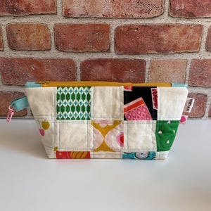 May include: A rectangular, patchwork fabric pouch with a yellow zipper and a light blue pull tab. The pouch features a variety of colorful fabric squares with patterns including green diamonds, pink shapes, and yellow flowers. The text "SEW CAERA" is visible on a small tag.