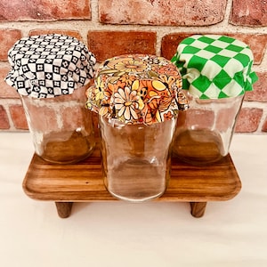 Op de afbeelding: Drie glazen potten met stoffen deksels. De deksels zijn in een zwart-wit geruit patroon, een bloemenpatroon en een groen-wit geruit patroon. De potten staan op een houten dienblad met poten.