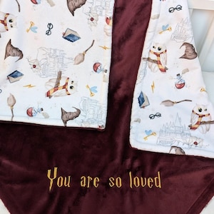 Puede incluir: Una manta suave con un lado blanco estampado con lechuzas, sombreros de mago, escobas, gafas y castillos, y un lado de color burdeos sólido. El lado burdeos presenta el texto amarillo bordado "You are so loved" en una fuente con serifa.
