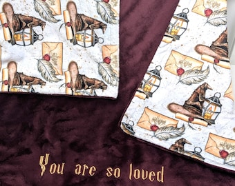 May include: A burgundy fleece baby blanket with a white cotton fabric panel featuring a repeating pattern of envelopes, feathers, and lanterns. The cotton panel has the text "You are so loved" embroidered in gold thread.