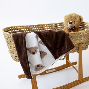 Può includere: Un lettino in vimini marrone con base a dondolo, una coperta marrone e bianca con facce di orso e un orsacchiotto di peluche marrone seduto all'interno.