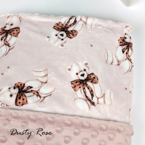 Pode incluir: Um close-up de um cobertor de bebê rosa empoeirado com um padrão de ursinhos brancos com laços marrons de bolinhas. A borda inferior tem uma borda texturizada com bolinhas. As palavras "Dusty Rose" são impressas na parte inferior.