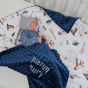 Peut inclure: Une couverture pour bébé bleu et blanche sur le thème d'Harry Potter. La couverture a un côté bleu marine avec le texte "Little Wizard" brodé dessus. L'autre côté de la couverture est blanc avec un motif de hiboux, de baguettes magiques et d'autres objets sur le thème d'Harry Potter.