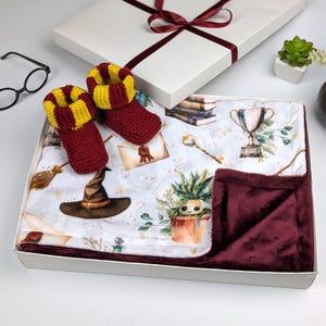 May include: A white gift box with a burgundy ribbon and bow. The box contains a burgundy and yellow baby blanket with a magical theme, featuring a witch's hat, a golden key, a trophy, and a potted plant. Two burgundy and yellow knitted baby booties are also included.