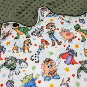 May include: A soft blanket featuring a white background with a colorful print of various animated characters including a space ranger, a cowboy, a cowgirl, a potato-shaped figure, and a green dinosaur, accented by small orange, yellow, and green stars.