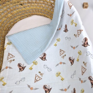 May include: A baby blanket with a light blue waffle-weave backing and a white patterned front. The front features repeating illustrations of a sorting hat, keys, glasses, and a golden snitch. The blanket is folded over a white surface.