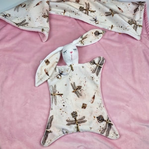 May include: A white bunny security blanket with a cream-colored fabric featuring a dragonfly and key pattern. The blanket is on a pink background.