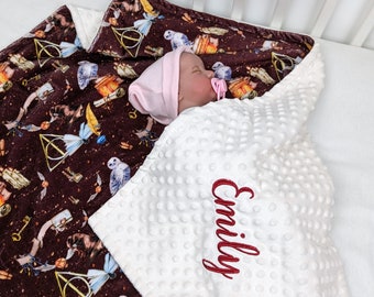 May include: A white baby blanket with a brown and gold Harry Potter themed print on one side and a white, textured side with the name "Emily" embroidered in red on the other side. A baby is sleeping on the blanket.
