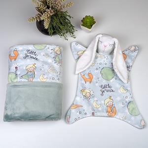 May include: A light blue baby blanket with a white bunny rabbit applique and a "Little Prince" theme. The blanket features a fox, stars, and planets.