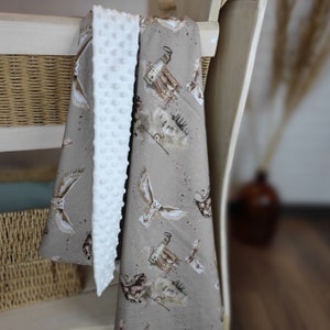 Op de afbeelding: Een babydeken met een bruin uilenprint en een witte, getextureerde achterkant. De deken heeft een herhalend patroon van uilen, flessen en brieven. De witte achterkant heeft een zachte, verhoogde punttextuur. De deken is over een lichtgekleurde houten constructie gedrapeerd.