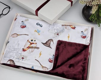 May include: A white and burgundy baby blanket with a Harry Potter theme. The blanket features a white side with a repeating pattern of owls, wands, hats, and the Hogwarts castle. The other side is a solid burgundy.
