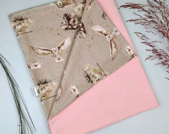 May include: A two-layered baby blanket. The top layer features a beige fabric with a repeating pattern of owls, letters, and other magical imagery. The bottom layer is a solid pink, textured fabric. A small white tag is visible on the top layer.