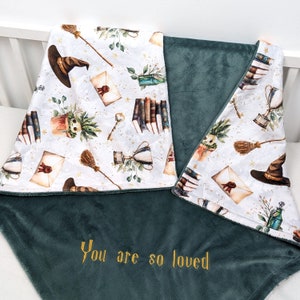 Könnte beinhalten: Eine doppelseitige Babydecke. Eine Seite hat einen weißen Hintergrund mit Aquarellillustrationen eines Hexenhuts, Büchern, einem Besen und einer Trophäe. Die andere Seite ist einfarbig dunkelgrün mit dem gestickten Text "You are so loved."