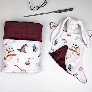 Può includere: Una coperta per bambini bianca e una coperta di sicurezza con bordo bordeaux. Le coperte presentano un motivo ripetuto di gufi, libri ed elementi magici. Sono presenti anche un paio di occhiali e una bacchetta.