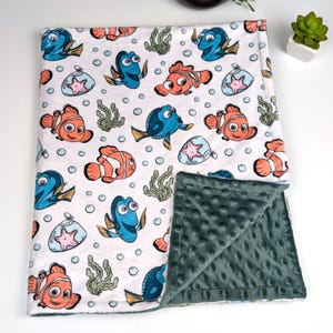 Può includere: Una coperta per bambini bianca con un motivo di pesci dei cartoni animati, alghe e bolle. La coperta ha un morbido retro verde scuro. I pesci sono arancioni e blu, con stelle marine in barattoli di vetro. La coperta è piegata.