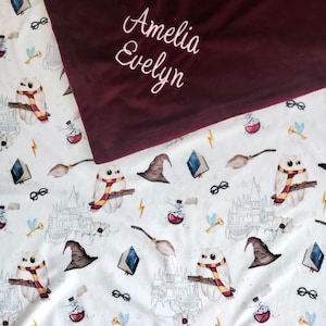 May include: A white fleece blanket with a burgundy fleece backing. The blanket features a repeating pattern of owls, wands, glasses, and a castle. The burgundy side has the name "Amelia Evelyn" embroidered in white.