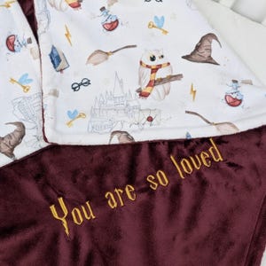 Può includere: Una copertina per neonati bianca e bordeaux a tema Harry Potter. Il lato bianco presenta un castello di Hogwarts, gufi e altri elementi magici. Il lato bordeaux ha il testo "You are so loved" ricamato in filo dorato.