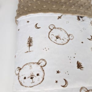 Op de afbeelding: Een zachte witte babydeken met een patroon van bruine berengezichten, maansikkels, sterren en dennenbomen. De bovenrand van de deken is afgewerkt met een gestructureerde bruine minky-stof met een verhoogd stippenpatroon voor een tactiel contrast.