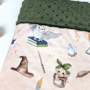 Puede incluir: Una manta para bebé con base rosa y borde verde. La manta presenta ilustraciones de un búho blanco, libros, un sombrero de mago, una varita, una botella de poción y una mandrágora. La manta está hecha de material suave.