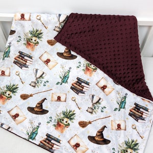 May include: A white and burgundy baby blanket with a whimsical pattern of witches hats, broomsticks, books, and potion bottles. The blanket is made of soft, plush fabric.