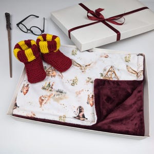 Könnte beinhalten: Ein Geschenkset mit weinroten und goldenen gestrickten Babyschuhen, einem Zauberstab und einer Brille. Das Set enthält eine weiße Decke mit weinrotem Plüschrand und eine weiße Geschenkbox, die mit einem weinroten Band gebunden ist.