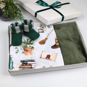 May include: A white gift box with a green ribbon and bow. The box is open and contains a green and gray striped knitted baby bootie, a green velvet blanket, and a white fabric with a green and white pattern of magical objects.